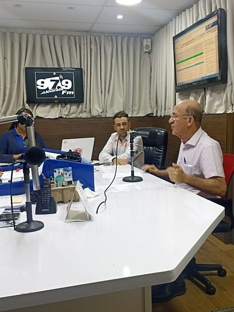 Deputado Federal Rubens Otoni usando camisa branca com uma TV ao fundo, ao lado de Paulo Antunes, presidente do PT de Rialma, trajando camiseta branca com uma placa da Rádio Alvorada ao fundo.