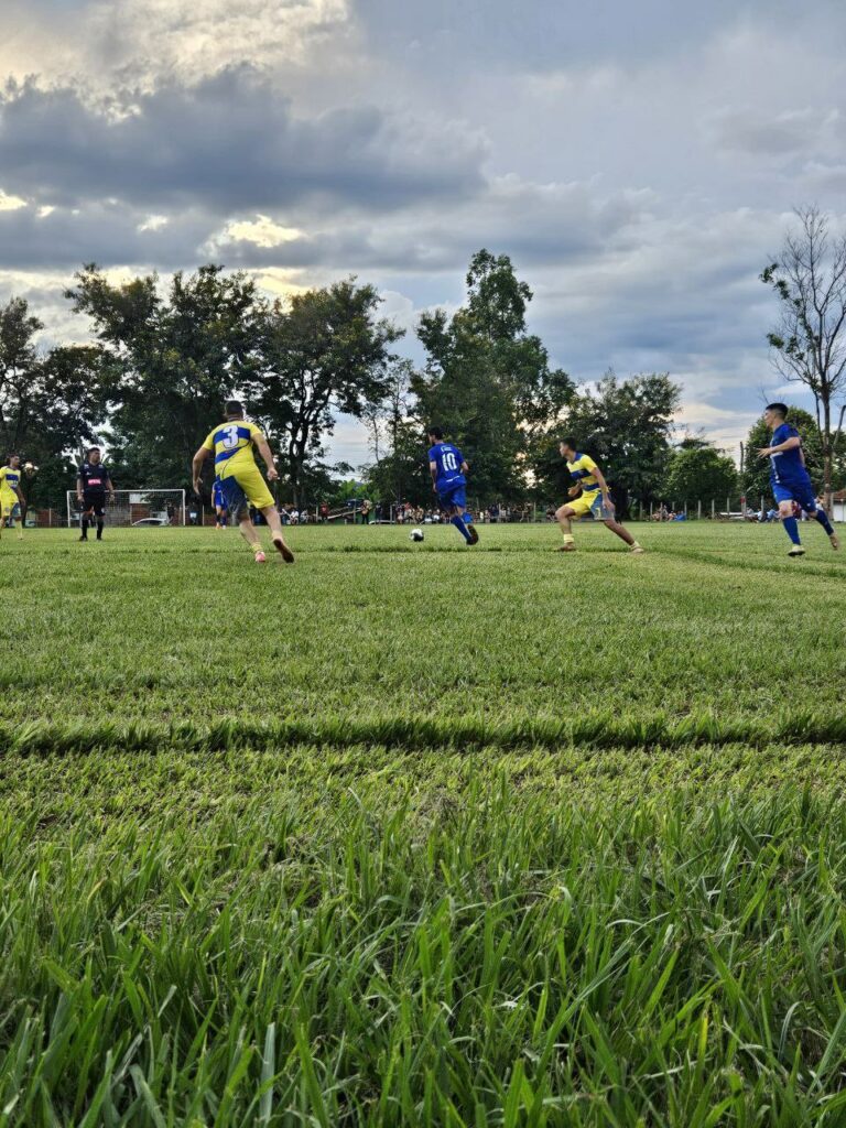Gramado verde com as equipes do Rissatti de azul e do Xixazão de amarelo disputando a bola branca